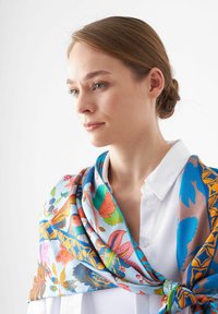 Foulard floral coloré en bleu, présentant des motifs complexes avec des oiseaux et des feuilles, drapé sur une chemise blanche à col. Texture de soie lisse.