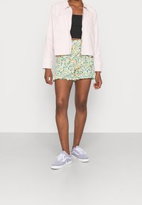 Ljusrosa skjorta med knappar, svart linne, gula blommiga shorts med volanger och lavendelfärgade sneakers med vita sulor. Enfärgad bakgrund.