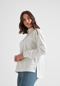 Woman wearing a white long-sleeve zip-up top with a small orange logo and blue fitted pants, standing against a plain light background.