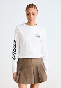 Bijela košulja dugih rukava sa malim logom na prsima i natpisom "Dickies" niz lijevu rukavu, u kombinaciji s smešom plisiranom suknjom.