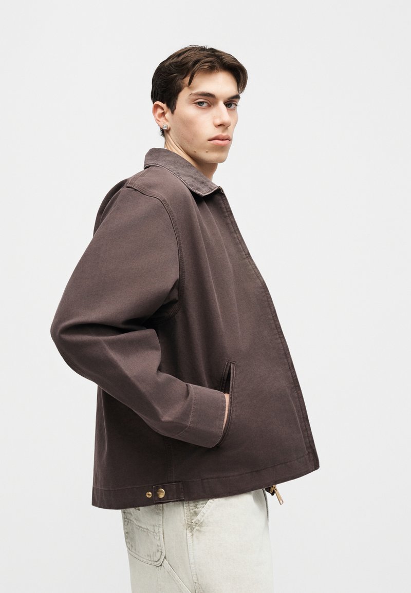 Hombre joven con una chaqueta marrón oscura y las manos en los bolsillos, mirando por encima del hombro sobre un fondo blanco liso.