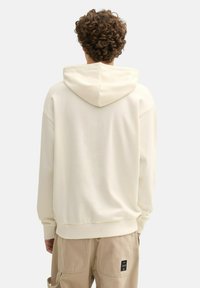 Sudadera color crema con capucha ajustable, puños y cintura de punto. Textura suave, diseño minimalista y etiqueta visible en pantalones beige.