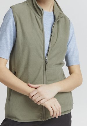 Osoba ubrana w jasnoszarą koszulkę z krótkim rękawem oraz oliwkowo zieloną kamizelkę polarową na zamek, ręce skrzyżowane przed talią.