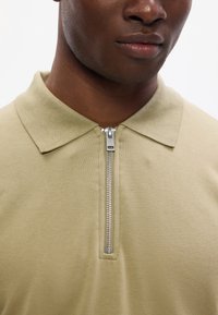 Khaki színű póló ing, cipzáros gallérral, puha tapintással és rövid ujjakkal. A cipzár ezüstfényű fémes bevonattal rendelkezik.