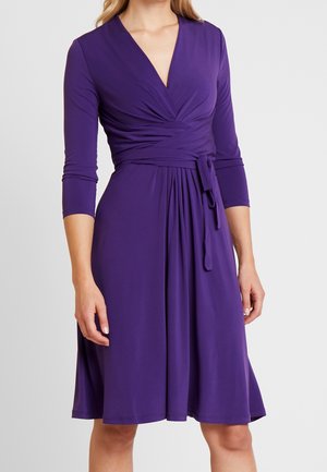 Kobieta ubrana w fioletową sukienkę kopertową do kolan z rękawami do łokcia i wiązaniem w talii.
