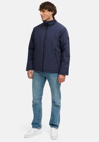 Tamsiai mėlynas zip-up džemperis su aukštu apykakle, dviem priekinėmis kišenėmis ir juodais akcentais. Dėvimas ant šviesiai mėlynų džinsų ir baltų sportinių batelių. Lygus paviršius.