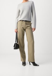 Nainen, jolla on päällään vaalea harmaa pitkähihainen paita, beige suora housu, musta vyö, mustat teräväkärkiset saappaat metallikärjillä ja musta käsilaukku.