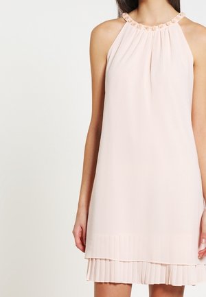 Ärmelloses hellrosa Kleid mit dekorativem Halsausschnitt und mehrlagigem plissiertem Saum, getragen von einer stehenden Person.