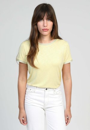 Femme aux cheveux longs bruns portant un t-shirt jaune clair à manches courtes et un pantalon blanc taille haute, debout devant un fond gris uni.