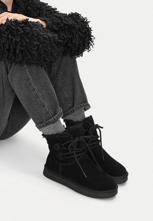 Persona seduta a gambe incrociate che indossa un maglione nero peloso, jeans scuri, calzini grigi e stivaletti in camoscio neri con fodera in pelliccia e lacci.