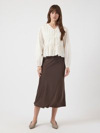 Kvinne står iført en kremfarget blouse med lange ermer og knytinger, samt et brunt skjørt til ankelen med beige lave hæler.