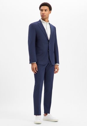 Man standing wearing a navy blue suit with a white striped shirt and white sneakers against a plain white background.