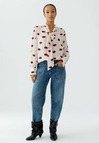 Weiße Bluse mit rotem Lippenprint, gebundenem Kragen und langen Ärmeln. Kombiniert mit hellblauen High-Waist-Jeans und schwarzen kniehohen Stiefeln.