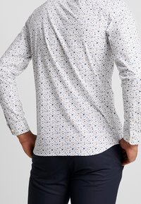 Biała koszula z długim rękawem, w drobny niebiesko-czerwony wzór kwiatowy, gładki materiał, slim fit, z guzikami przy mankietach i zaokrąglonym dołem.