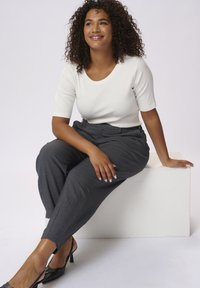 Pantaloni grigi su misura con dettagli sulle cuciture laterali, abbinati a un top bianco aderente a maniche corte. Modella seduta su un blocco bianco. Tacchi neri.