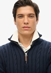 Marine kabelgebreide zip-up trui met hoge kraag over een wit shirt. Voorzien van een zilveren rits met een bruine leren accent.