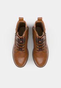 Par bruna läderstövletter med svarta snören, sidodragkedjor och dragflikar, sedd ovanifrån på en vit bakgrund.