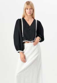 Blouse noire courte avec des manches ballons, dotée d'un décolleté en V, d'une fermeture à boutons à l'avant et d'une bordure en dentelle blanche. Associée à une jupe blanche à motifs.