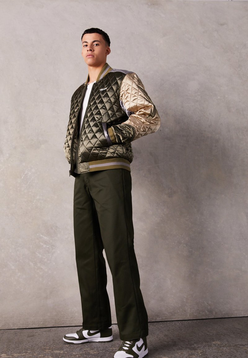 Veste bomber matelassée avec un corps vert olive et des manches à motifs beige clair, associée à un pantalon vert foncé et des baskets avec des accents blancs.