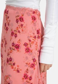 Ljusrosa blommig kjol med ett genomskinligt tyg, prydd med livfulla rosa och lila blommor samt gröna blad. Midjebandet har en delikat spetskant.