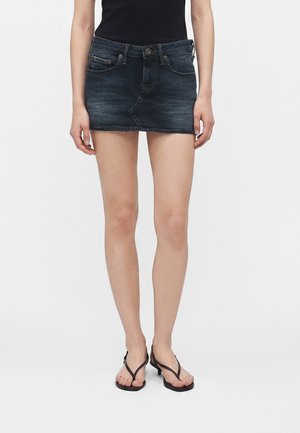 Persoon draagt een donker denim minirok, zwart mouwloos top en zwarte sandalen met hakken, staand tegen een effen witte achtergrond.