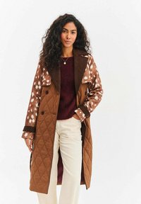 Brązowy, pikowany trencz z łatami w cętki i kontrastującym kołnierzem. Posiada dwurzędowe guziki i luźny fason. W połączeniu z bordowym swetrem i jasnobeżowymi spodniami.