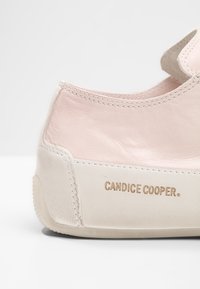 Gros plan d’un talon de basket en cuir rose pâle et blanc avec « CANDICE COOPER » embossé sur la semelle blanche.