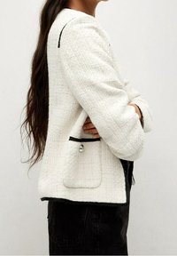 Veste blanche texturée avec bordure noire, dotée d'une poche avant et d'une coupe classique. Le tissu semble tissé avec un léger éclat.