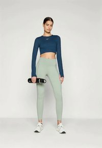 Crop top sportiv de culoare albastră închisă, pantaloni leggings verzi deschis cu talie înaltă și adidași albi. Modelul ține o sticlă de apă neagră, stil minimalist.