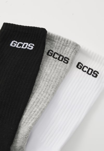 Trois paires de chaussettes côtelées en noir, gris et blanc, chacune arborant le logo de la marque "GCDS" dans des couleurs contrastées près de la partie supérieure.