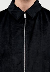 Giacca nera con zip in velluto, con colletto a risvolto e cerniera argentata davanti. Texture liscia.