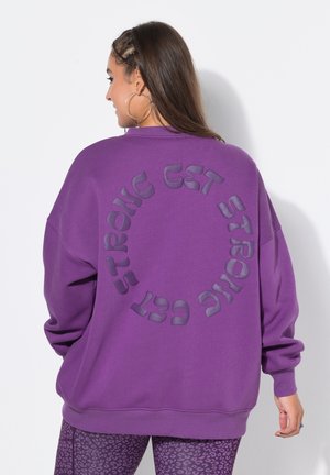 Kobieta o długich brązowych włosach, ubrana w fioletową, oversize'ową bluzę z napisem „GET STRONG” w okrągłym wzorze na plecach oraz fioletowe legginsy w wzory.