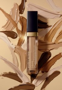 Estée Lauder Futurist Soft Touch Brightening Skincare i en klar tube med svart lock. Bakgrunden visar olika nyanser av beige och brun foundation.
