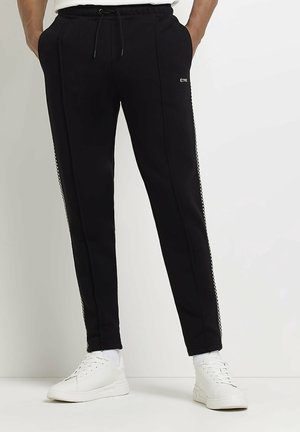 Partie inférieure portant un pantalon de jogging noir avec des bandes à carreaux sur les côtés et des baskets blanches, mains dans les poches.