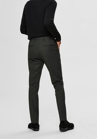 Dark green tailored pants with slim fit, five-pocket design, and belt loops, made from a smooth, textured fabric. Black footwear worn.