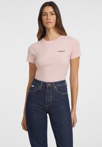 Gaišs rozā pieguļošs t-krekls ar īsām piedurknēm un apaļu kakla izgriezumu, featuring neliels "GUESS JEANS" logo tumšā krāsā. Saskaņots ar tumši ziliem augstās jostas iešūtiem džinsiem.