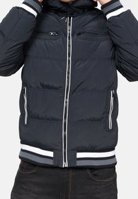 Tummansininen topattu takki, jossa on etuvetoketju, kaksi rintataskua, valkoiset yksityiskohdat sekä raidalliset joustavat hihansuut ja helma. Pintakuvioitu viimeistely.