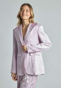 Blazer viola chiaro con finitura testurizzata, colletto a punte, tasche frontali e dettaglio con bottoni sulle maniche, indossato sopra pantaloni coordinati.