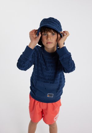 Sweatshirt texturé bleu marine avec motif de logo et poche, associé à un short corail avec un motif blanc. Enfant portant un chapeau en seau assorti.