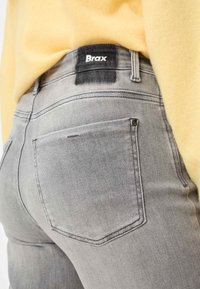 I jeans di jeans grigi presentano una vita alta, tasche posteriori e una toppa con il logo Brax sulla vita, abbinati a un maglione giallo.
