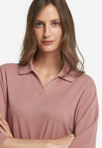 Ljusrosa polo tröja med korta ärmar, med en traditionell krage och en texturerad yta. Tyget verkar mjukt och något stretchigt.