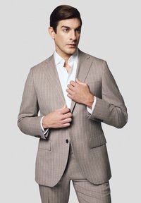 Jasnobrązowy garnitur w prążki wykonany z gładkiego materiału. Posiada dwa guziki, klapy z wycięciem i dopasowany krój. W połączeniu z białą koszulą elegancką.