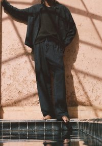 Zwarte loszittende blouse en broek, gemaakt van zachte stof, met een casual ontwerp. Model staat blote voeten bij een zwembad tegen een gemarmerde muur.