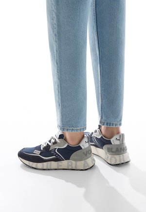 Person iført lyseblå jeans og marineblå, grå og hvide sneakers med tykke såler, stående på en hvid overflade.