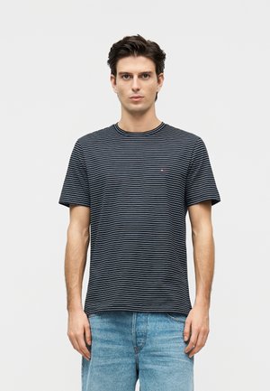 Mand med kort mørkt hår iført en sort og hvid stribet t-shirt og lyseblå jeans, stående foran en ensfarvet hvid baggrund.