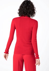 Rood, passend langemouwen top met een hoge hals en gerimpelde manchetten. Zachte textuur met een gladde afwerking. Gecombineerd met bijpassende rode broek.