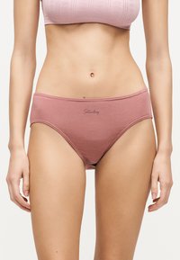 Γυναίκα που φοράει μωβ υψηλοκάβαλα εσώρουχα με το κείμενο "Saturday" μπροστά και ένα αντίστοιχο υφασμάτινο μωβ τοπ, στέκεται μπροστά σε λευκό φόντο.