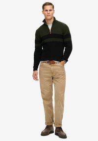 Sweter z dzianiny z ćwierć zamkiem, w ciemnozielone i czarne paski. Połączony z beżowymi spodniami i brązowymi butami na sznurowadła.