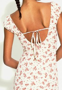 Kukka-aiheinen minimekko, jossa on kermaantuoteinen pohja, ribbitekstuuri, puffihihat ja matala selkä siteellä. Pieniä punaisia ja vaaleanpunaisia kukkia ympäriinsä.