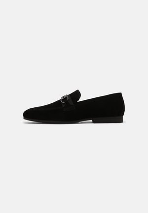 Zwarte suède loafer met metalen bit-detail, lage hak en instapmodel, zijaanzicht op een effen achtergrond.